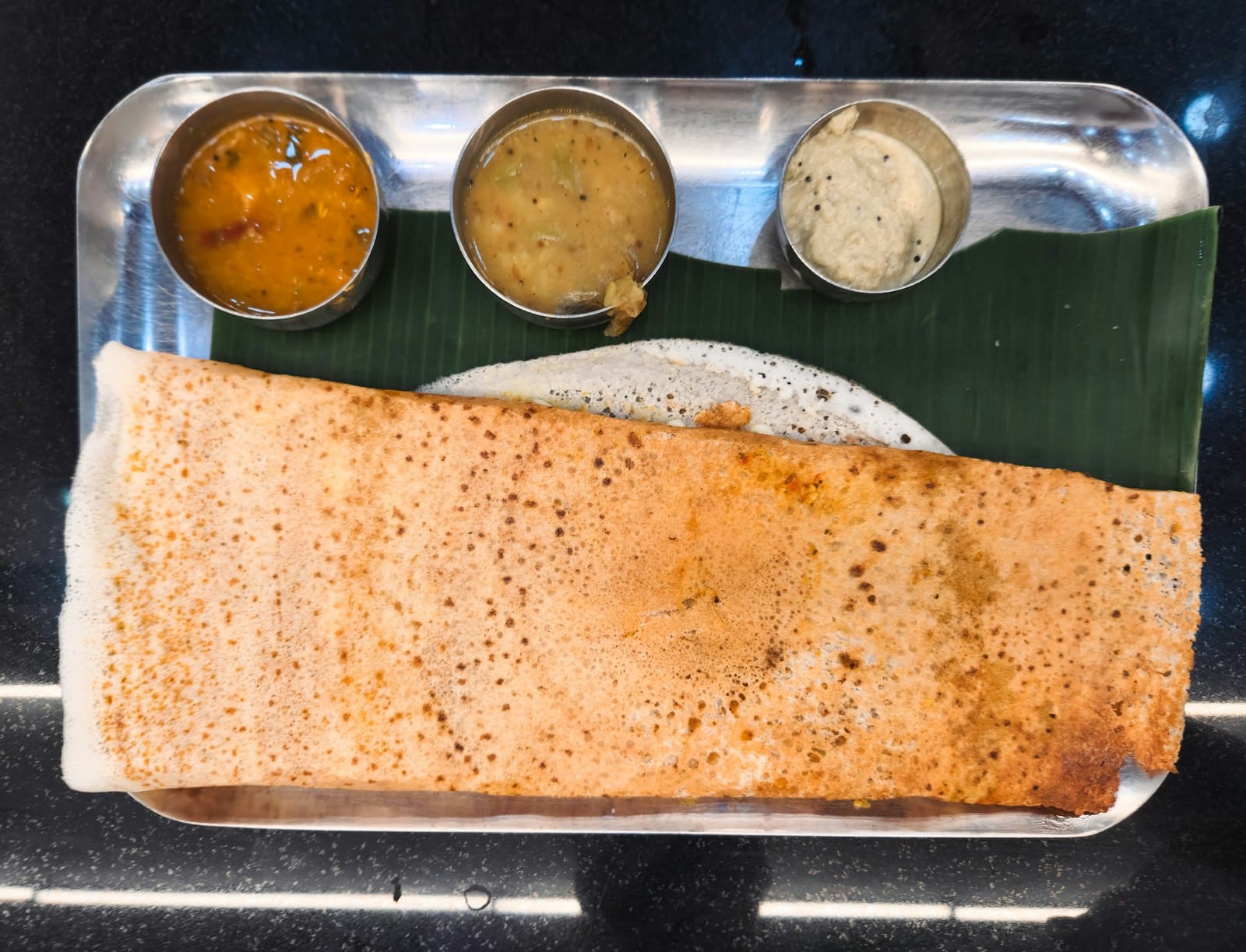 A large dosa on a steel tray with three chutneys
